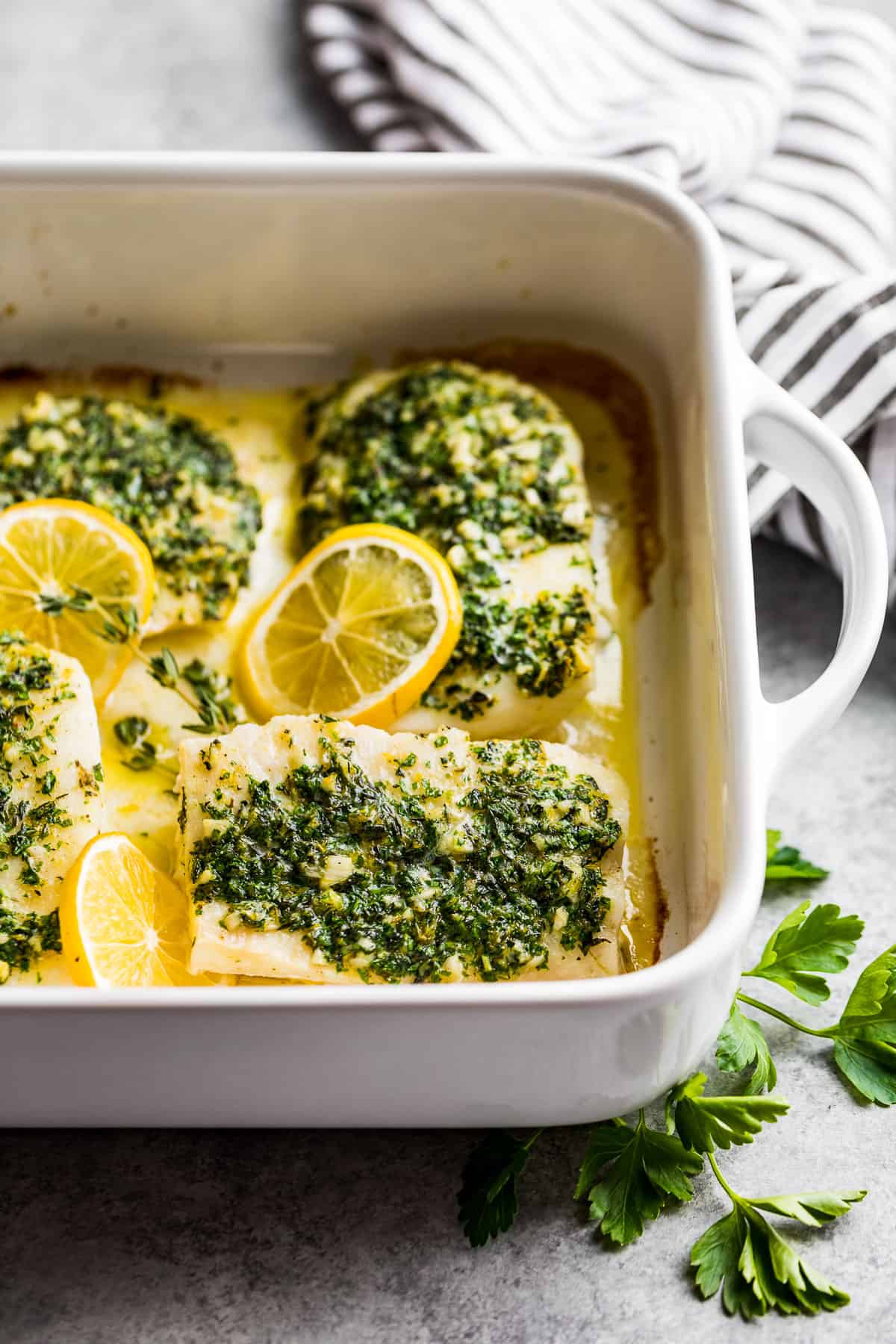 A side view of Oven Baked Lemon Herb Cod in a white baking dish with parsley sprigs and a striped linen near the dish.