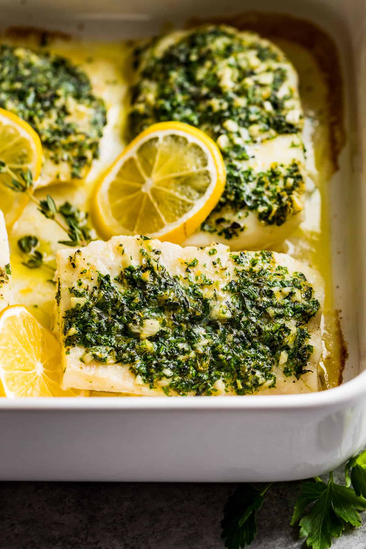 A side view of Oven Baked Lemon Herb Cod in a white baking dish with parsley sprigs and a striped linen near the dish.