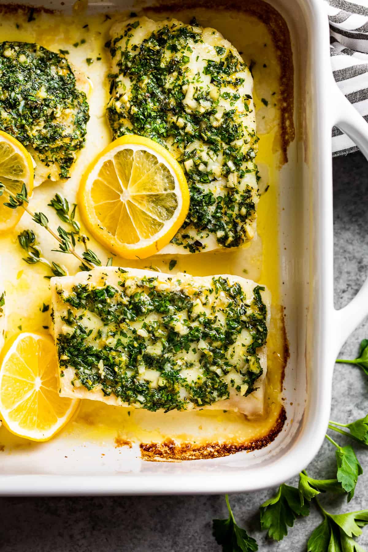 A large white baking dish with Oven Baked Lemon Cod with a striped linen and lemon slices around the dish.