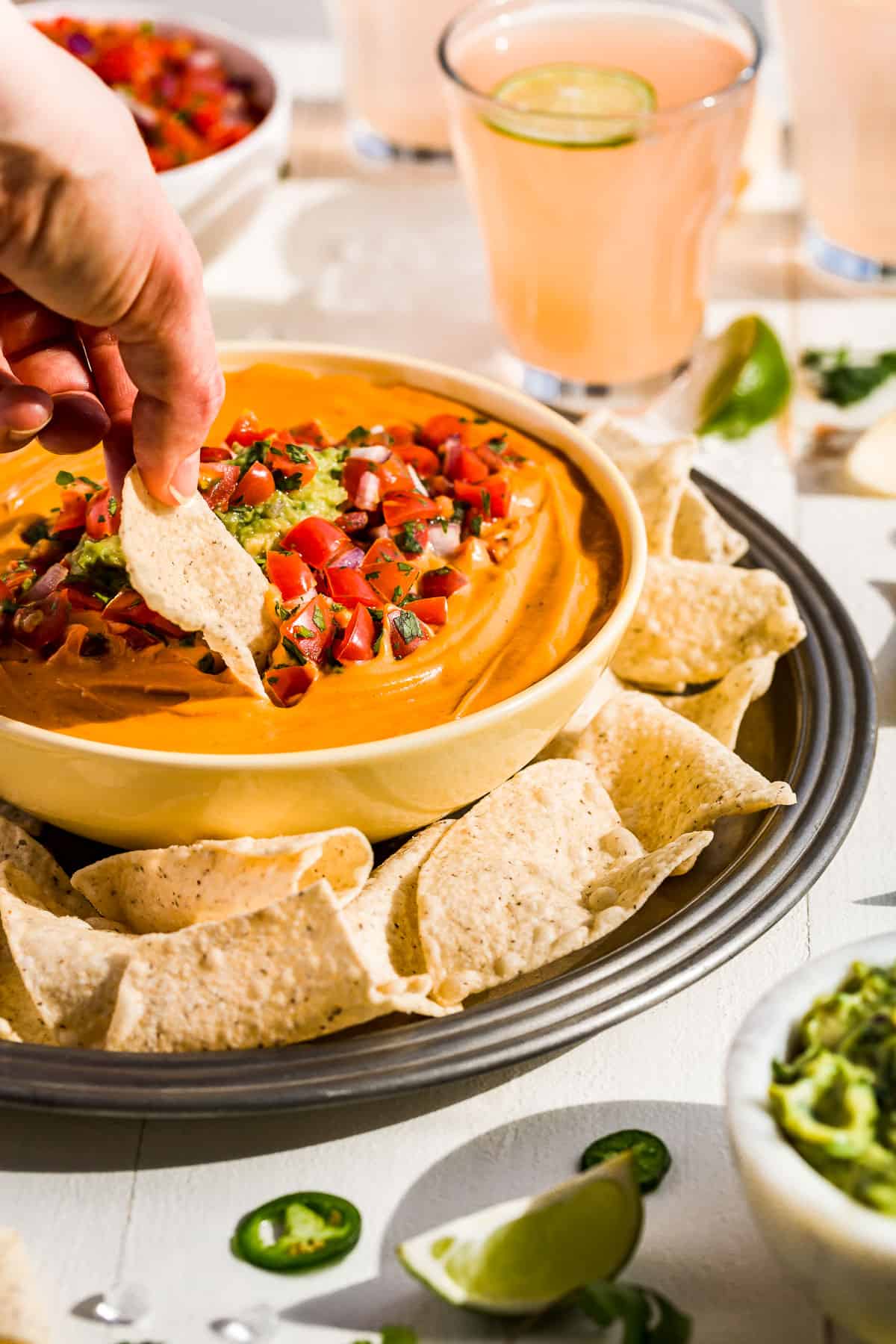 A hand dipping a corn chip into the bowl of vegan queso surrounded by chips with drinks in the background.