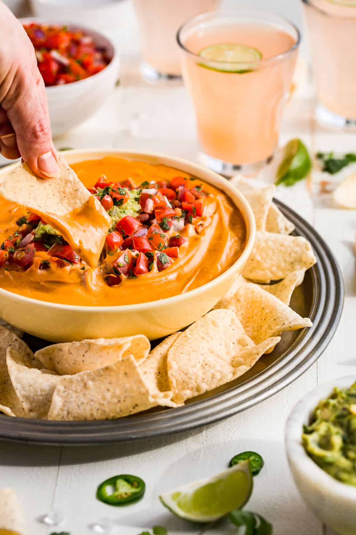 A hand dipping a chip into the bowl of queso with drinks in the background.
