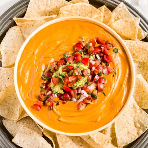 Vegan Queso in a bowl topped with Pico de Gallo and Guacamole surrounded by chips.