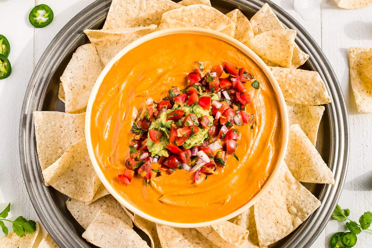 Vegan Queso in a bowl topped with Pico de Gallo and Guacamole surrounded by chips.