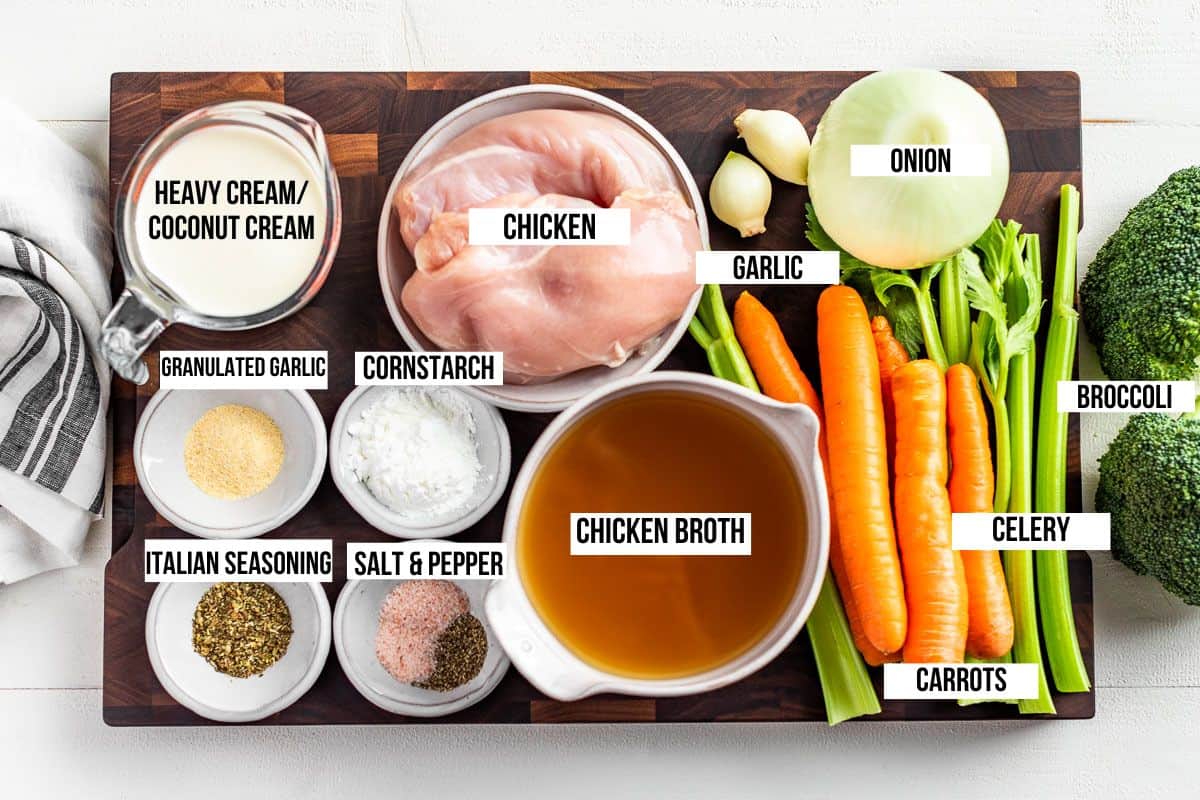 Chicken, chicken broth, carrots, onion, garlic, celery, broccoli, spices, and cream in bowls on a wood cutting board.