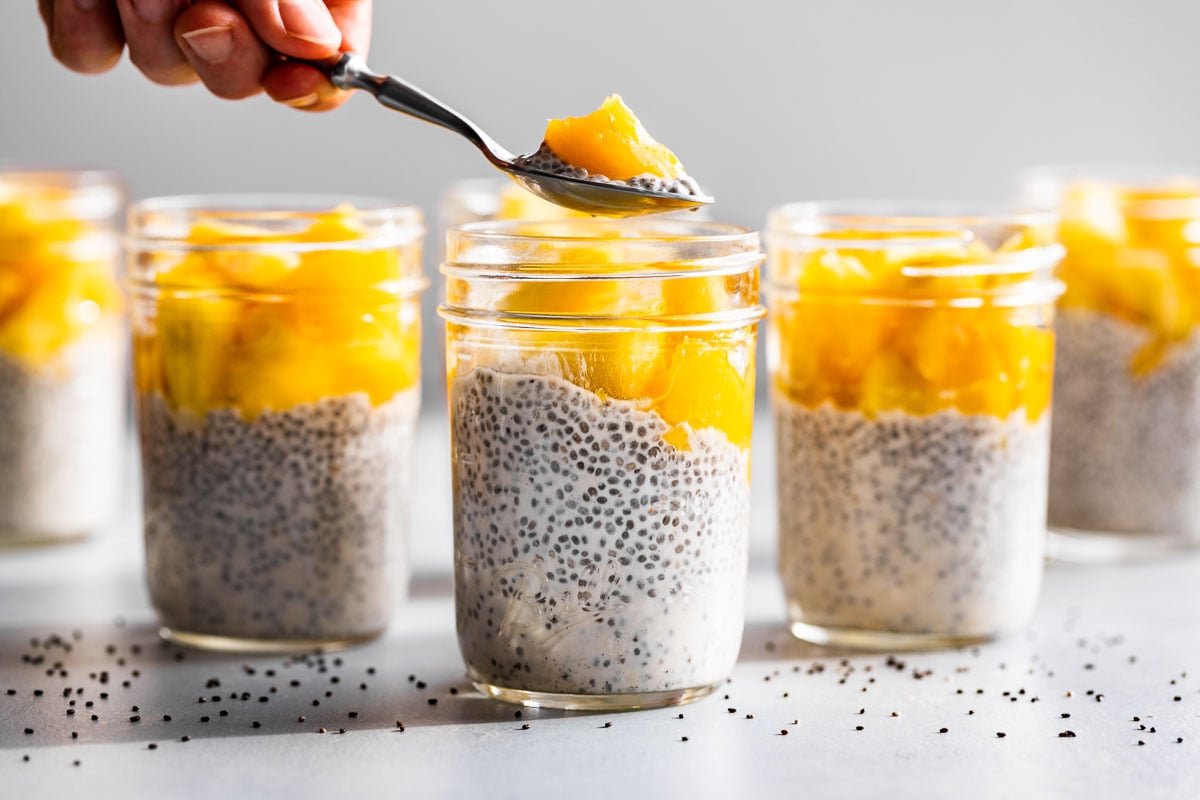 5 jars of Creamy Coconut Chia Pudding topped with pineapples and mangoes with a spoonful being taken out.