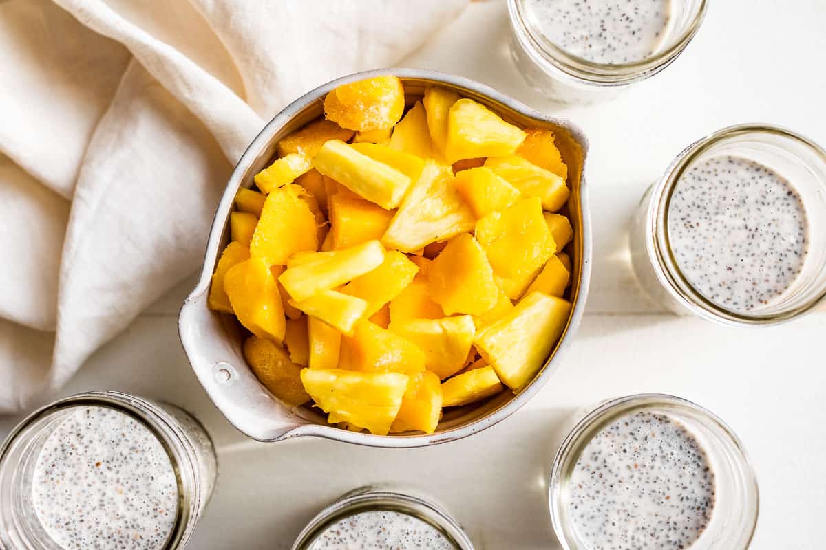 A bowl of pineapple and mango with coconut chia pudding jars around it.