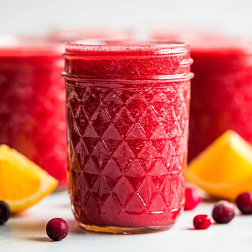 Three glasses of cranberry orange smoothie with frozen cranberries and orange wedges placed around them.