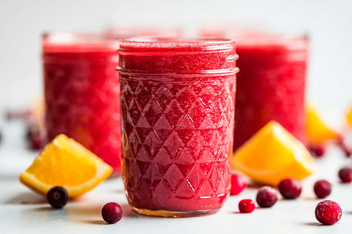 Three glasses of cranberry orange smoothie with frozen cranberries and orange wedges placed around them.
