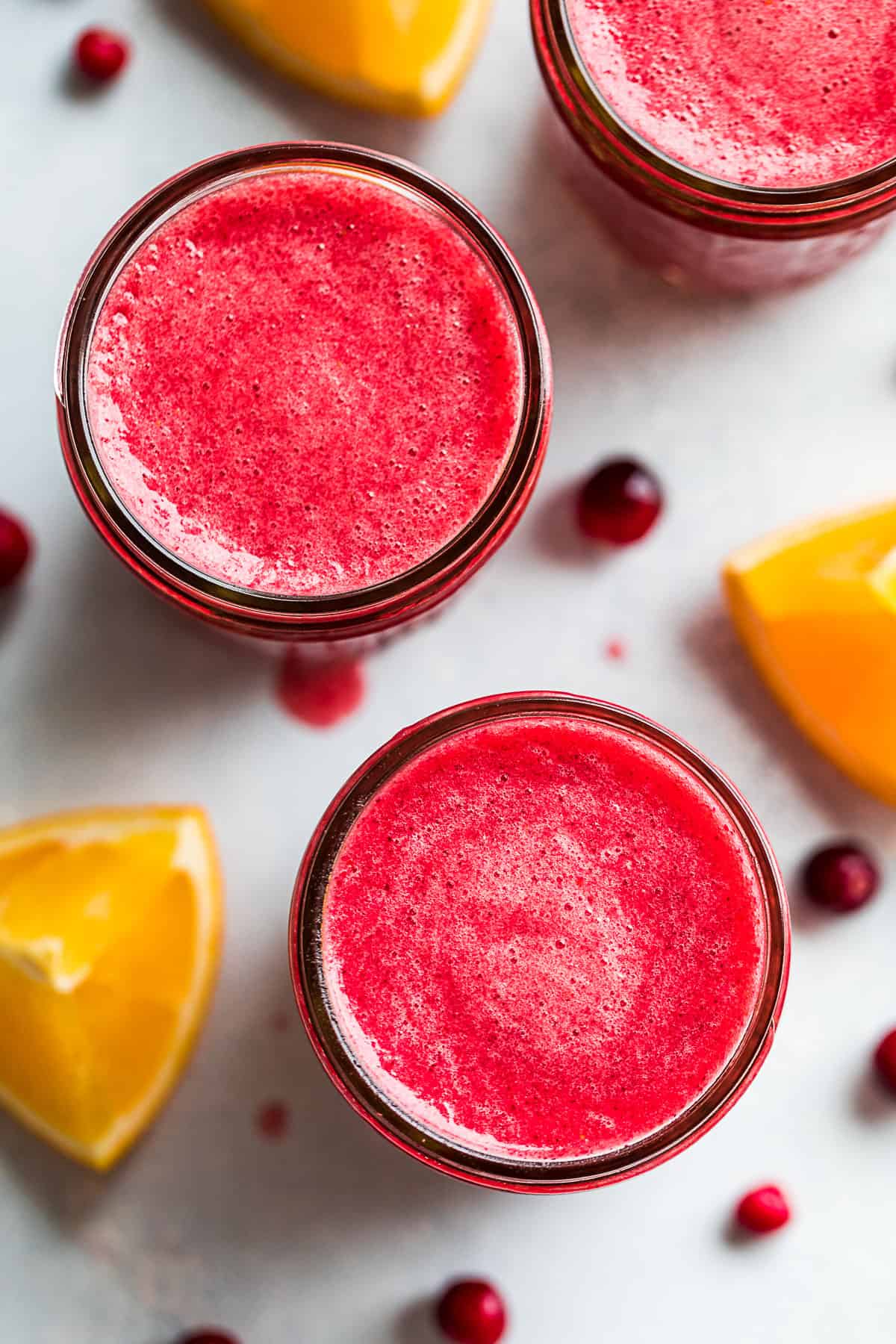 Straight down view of Cranberry Smoothie poured into 3 glasses with cranberries and orange wedges around them.