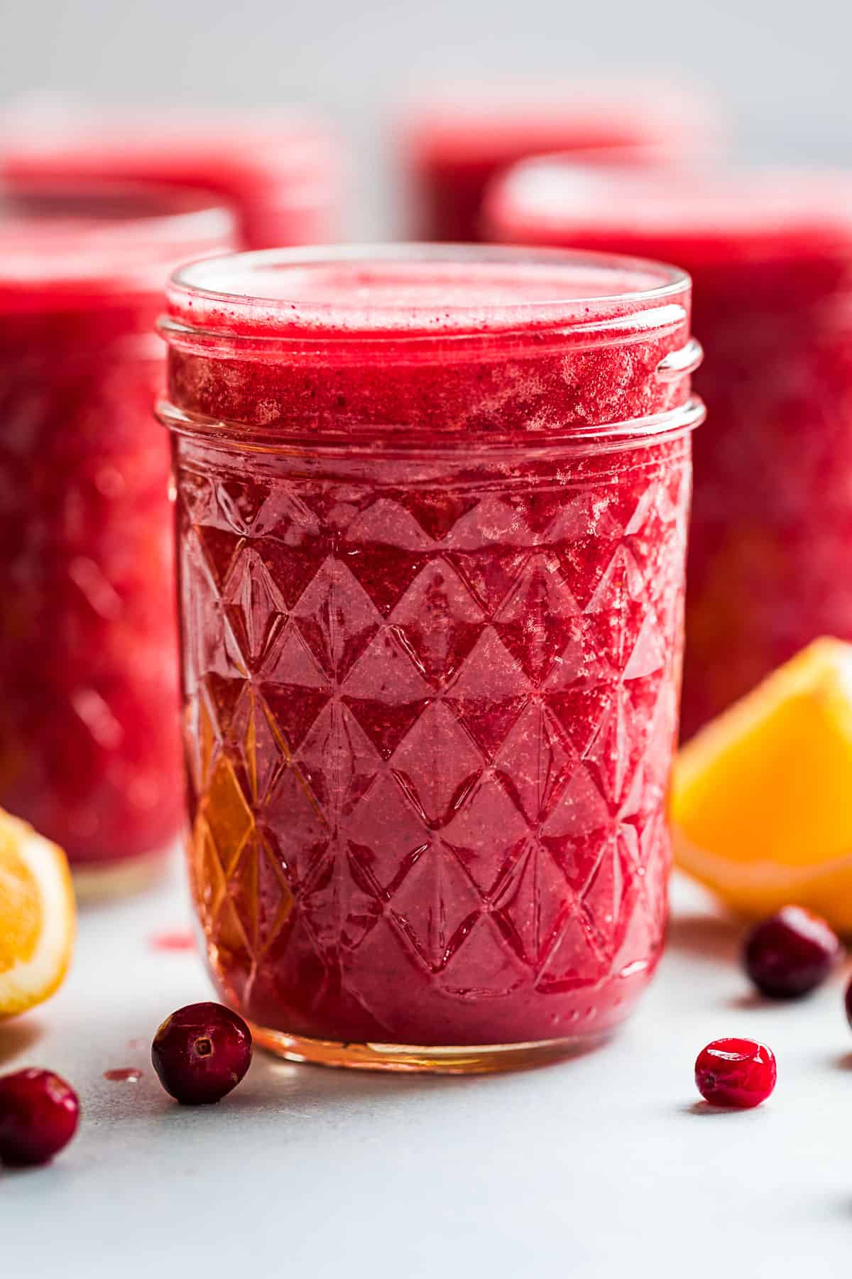 Backlit glasses filled with Cranberry Smoothie with frozen cranberries and orange wedges around them.