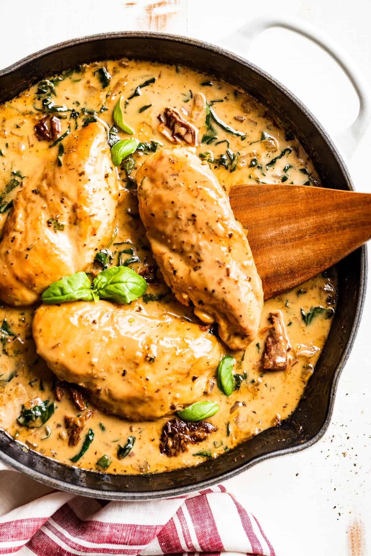 Creamy Tuscan Chicken in white skillet with basil leaves, a wooden serving spoon, and red striped linen.