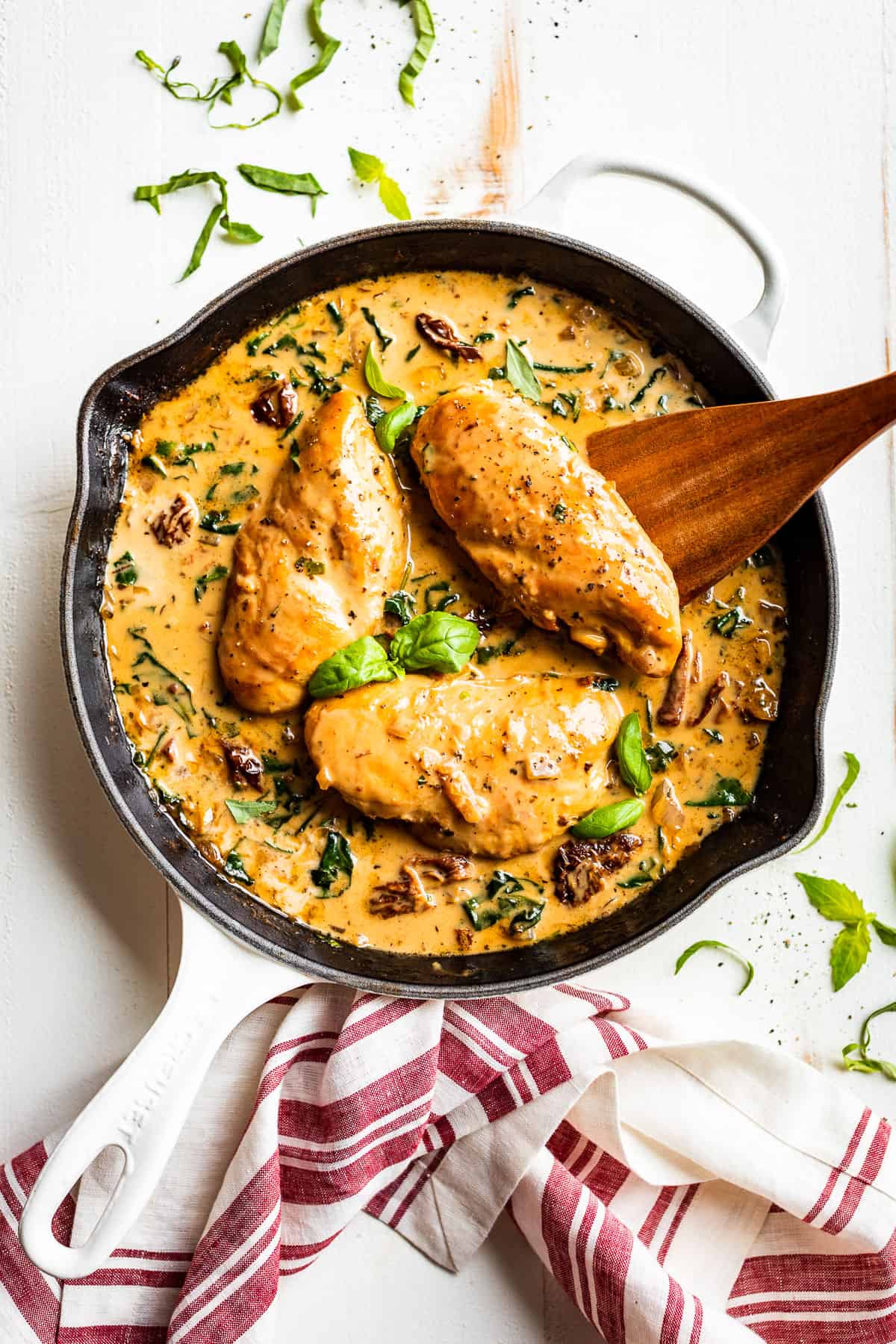 Creamy Tuscan Chicken in white skillet with basil leaves, a wooden serving spoon, and red striped linen.