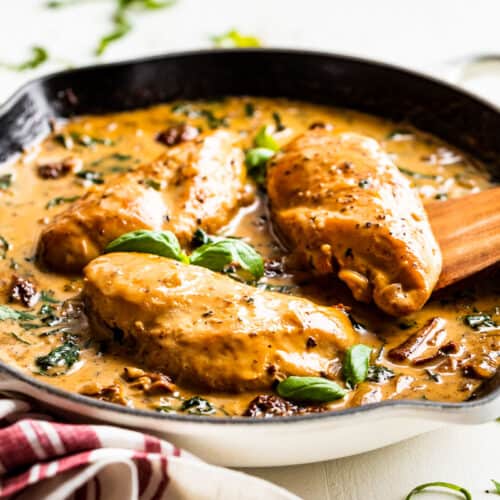 Creamy Tuscan Chicken in a white skillet with a red and white linen and a wooden serving spoon.
