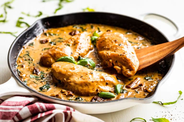Creamy Tuscan Chicken in a white skillet with a red and white linen and a wooden serving spoon.