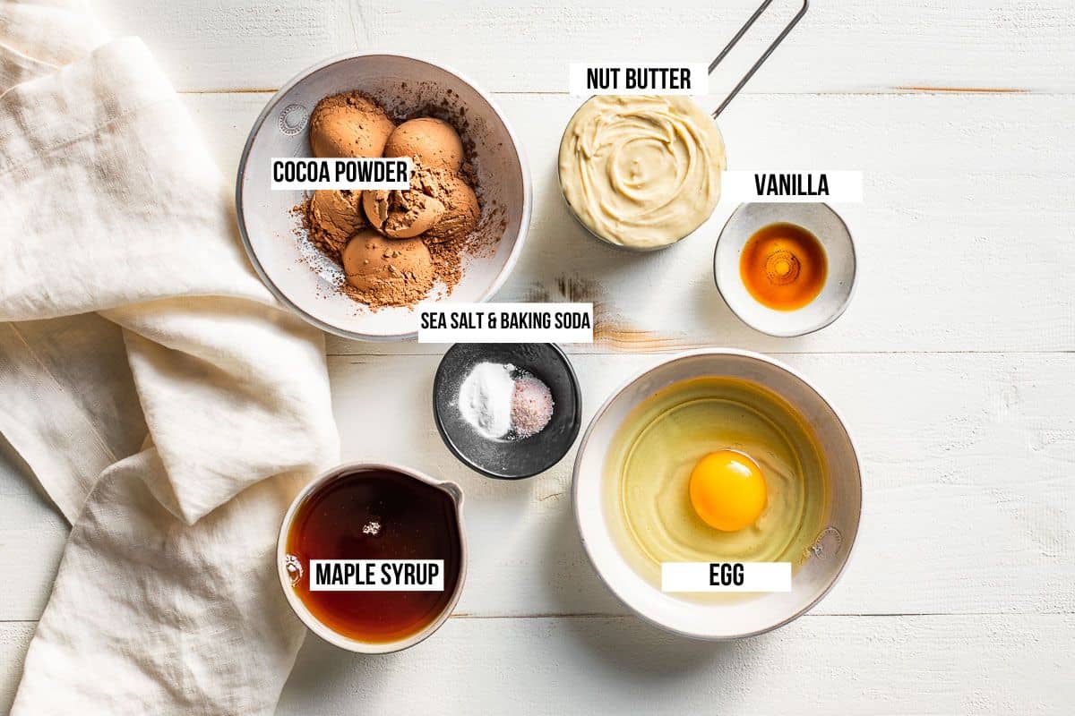 Cocoa powder, maple syrup, nut butter, baking soda, egg, vanilla and sea salt in small bowls on a white background.