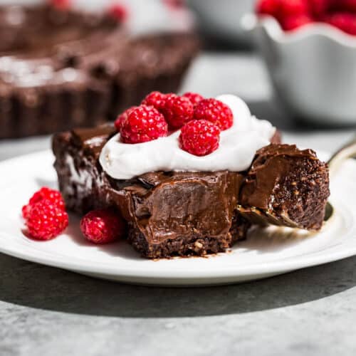 A slice of chocolate tart topped with whipped cream and raspberries with a fork scooping a bite off the tip of the slice.