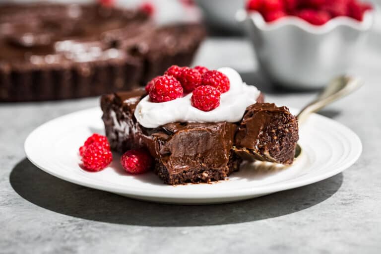 A slice of chocolate tart topped with whipped cream and raspberries with a fork scooping a bite off the tip of the slice.