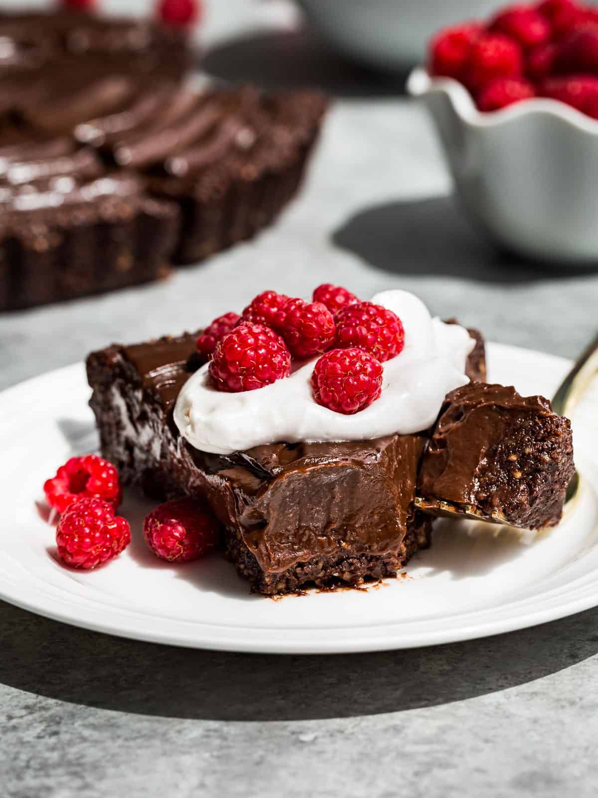 Close up view of a slice of Healthy Chocolate Tart topped with whipped cream and raspberries with a bite cut off the front with a fork.