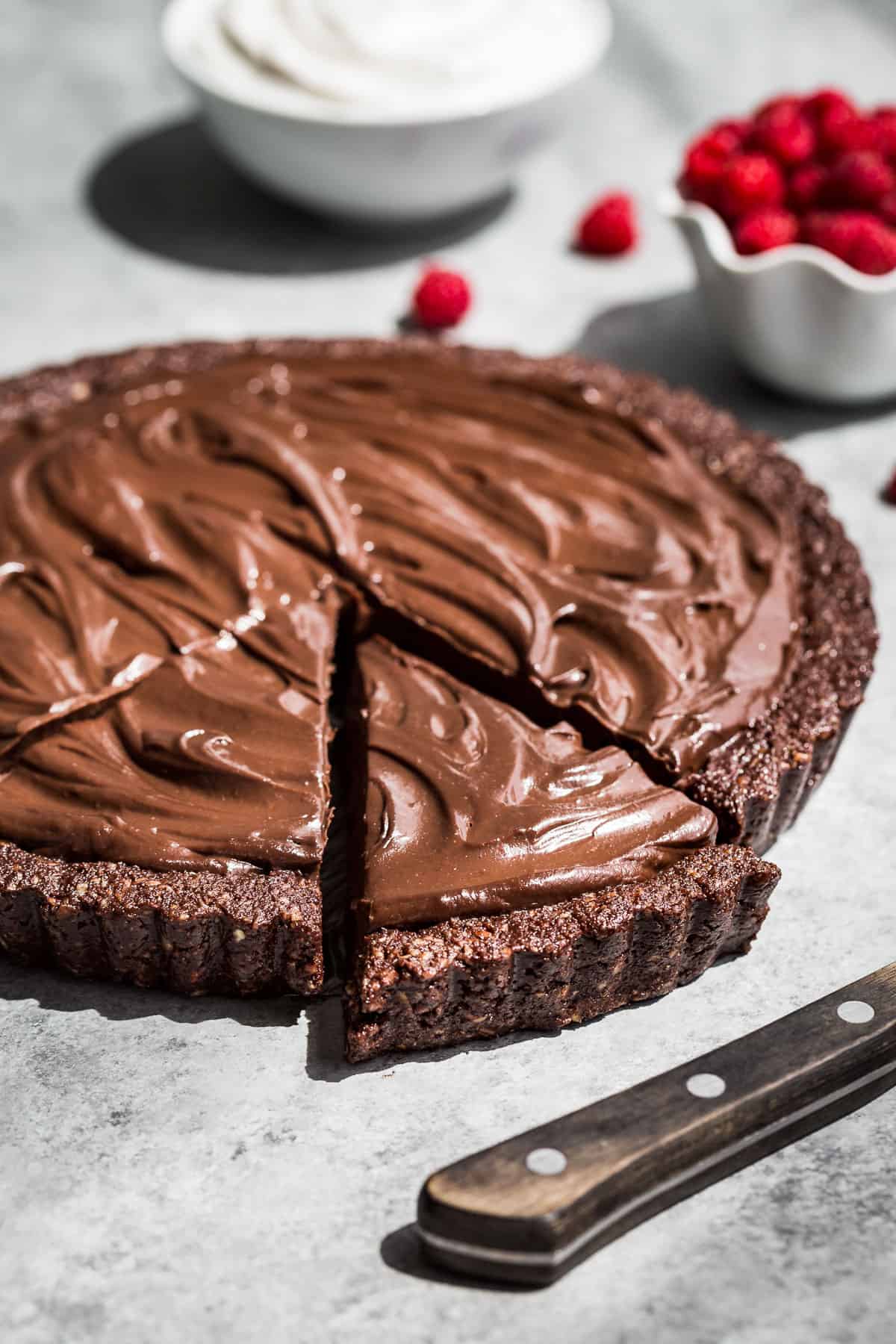 The whole Healthy Chocolate Tart with 2 slices cut and bowls of whipped cream and raspberries in the background.