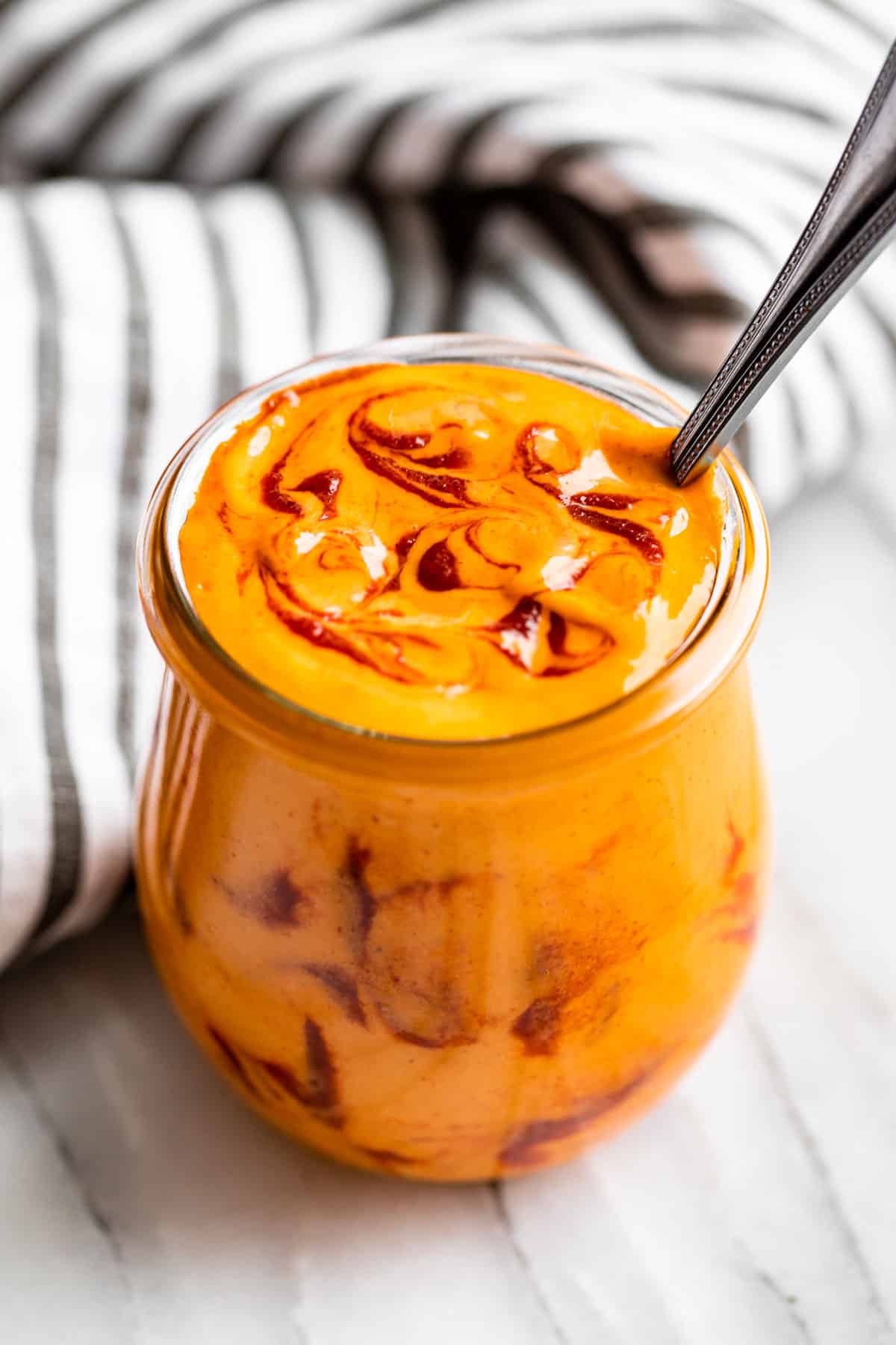 Side view of Sriracha Mayo in a glass jar with a silver spoon in it with sriracha swirled on top.