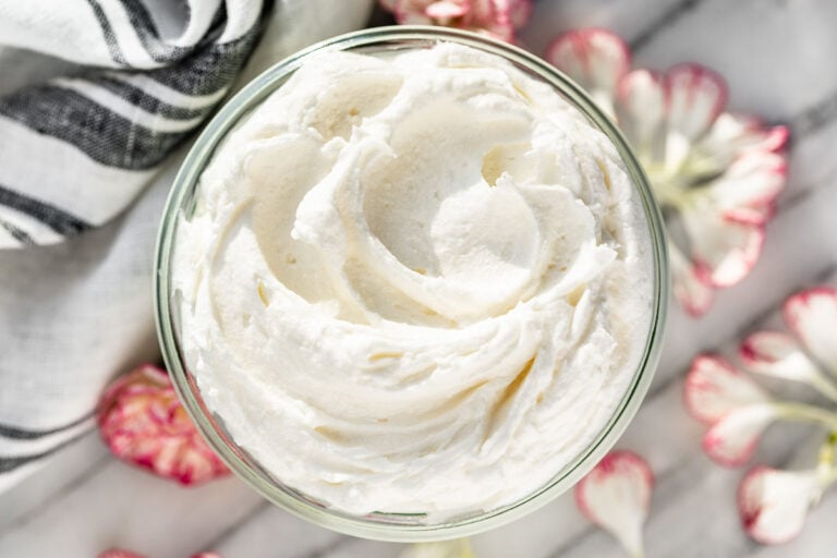 Whipped Body Butter in a glass jar with a grey linen in the background and flower petals surrounding it.