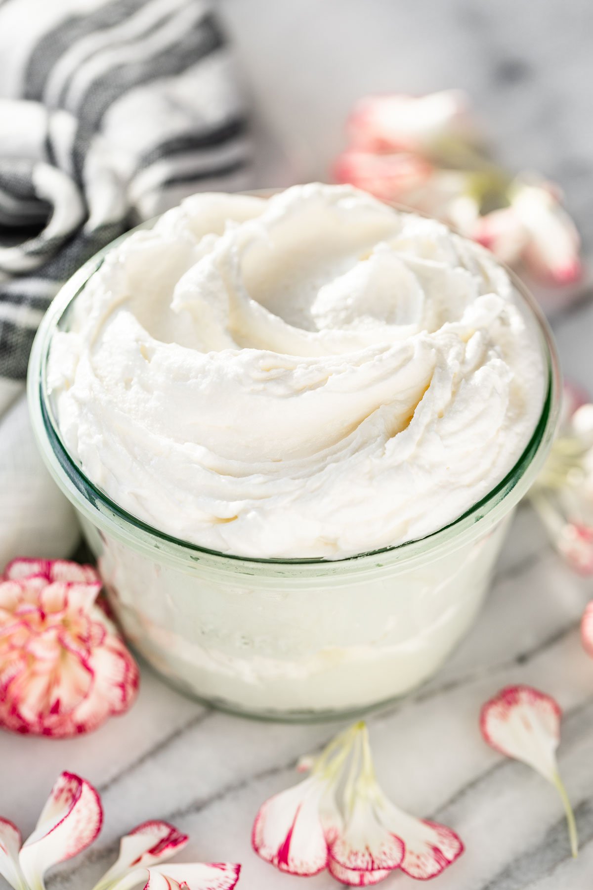 Side view of Whipped Body Butter in a glass jar with a grey linen in the background and flower petals surrounding it.