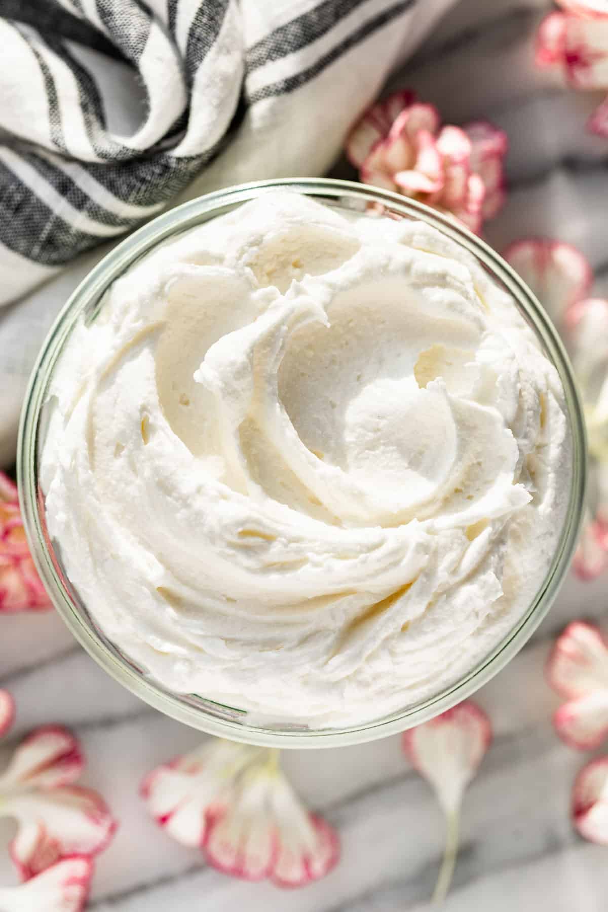 Straight down view of Whipped Body Butter in a glass jar with a grey linen in the background and flower petals surrounding it.