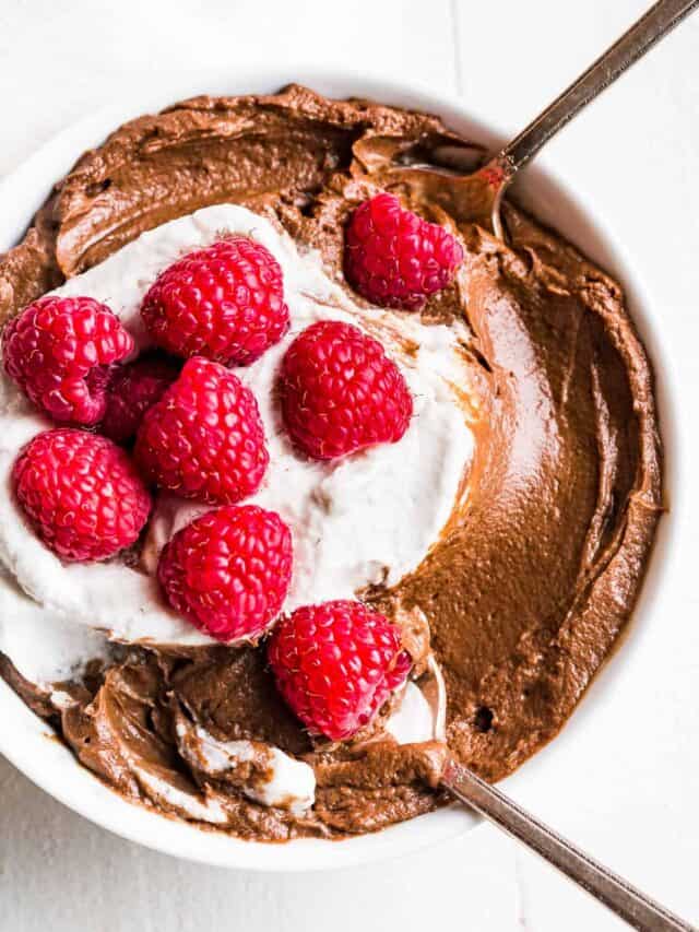 Straight down view of Avocado Chocolate Mousse in a white bowl topped with whipped cream and raspberries with 2 spoons in it.