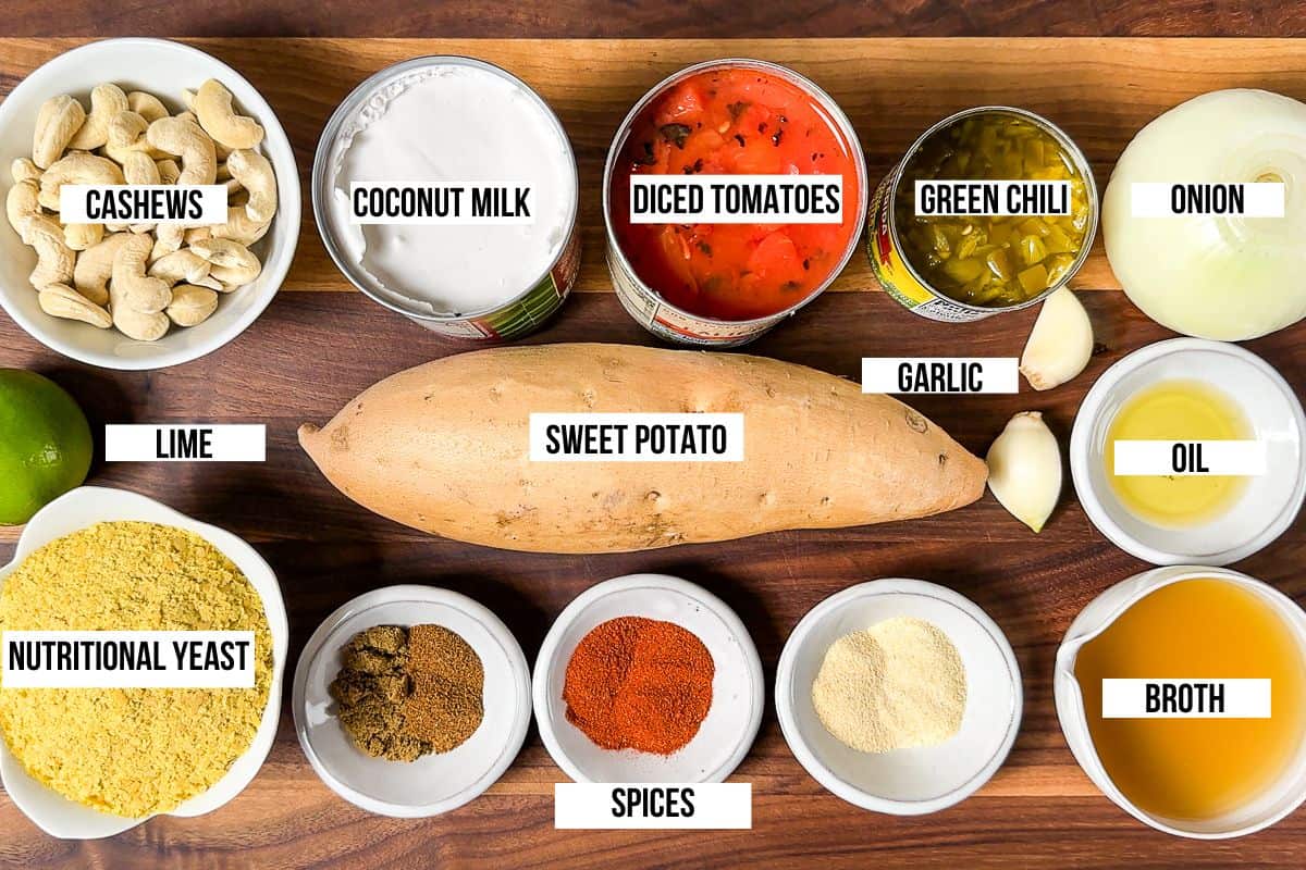 Cashews, nutritional yeast, sweet potato, coconut milk, spices, tomatoes, green chili, onion, and garlic on a wood cutting board.