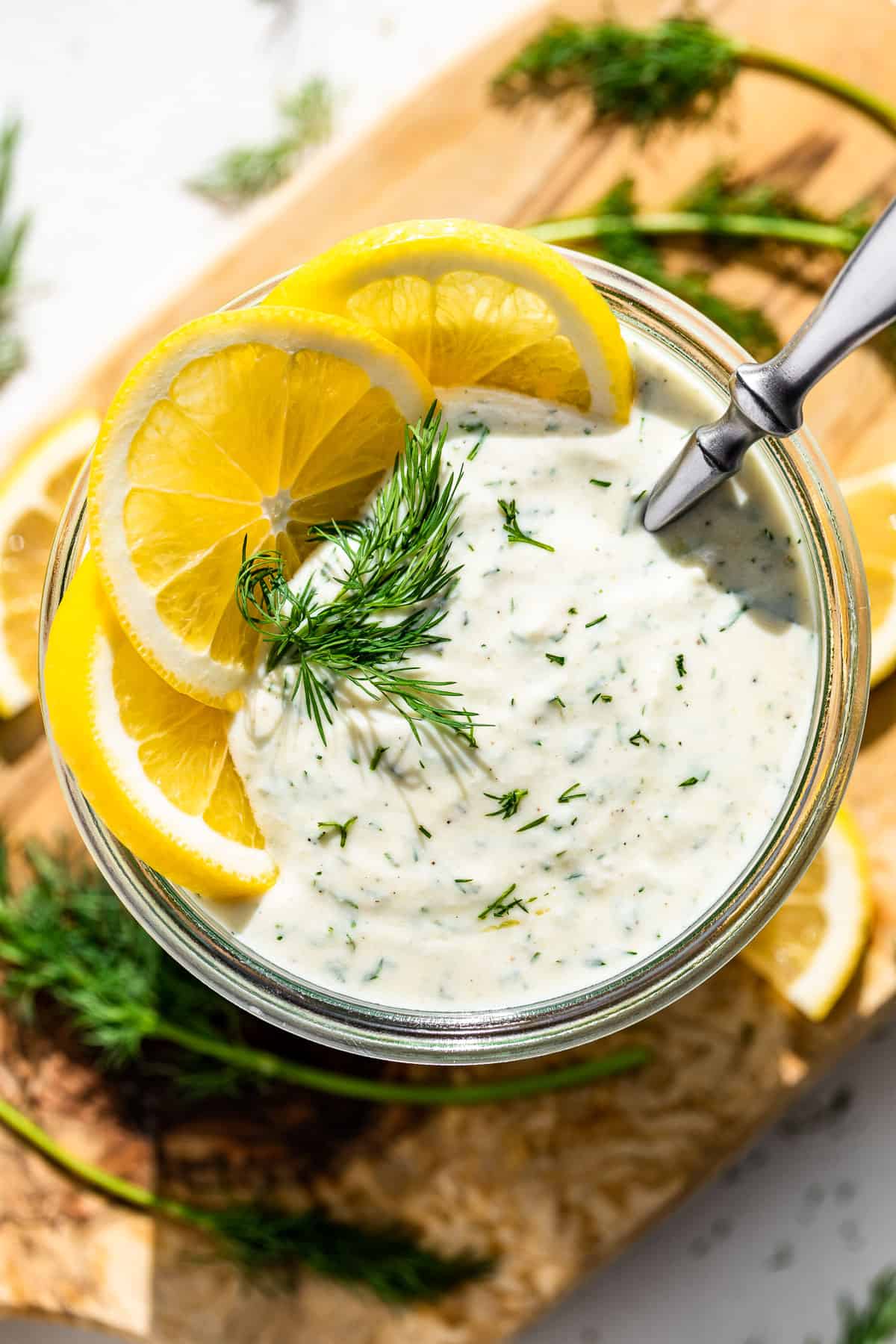 Straight down view of Greek Yogurt Dressing in a clear glass jar topped with lemon slices and a sprig of dill set on a wood cutting board.