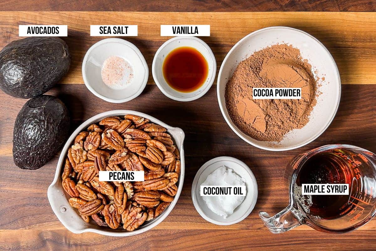 Avocados, cocoa powder, vanilla extract, maple syrup, coconut oil, and pecans in small bowls on a wood cutting board.