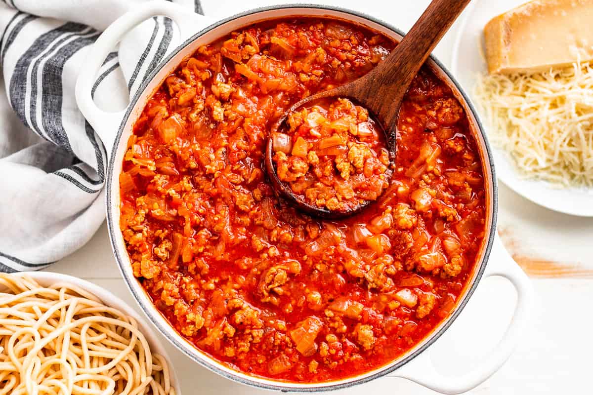 Finished Homemade Spaghetti Sauce in a white pot with a wood ladle, with bowls of cooked spaghetti and grated parmesan on the side.