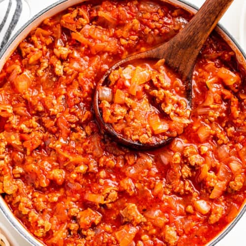 Finished Homemade Spaghetti Sauce in a white pot with a wood ladle, with bowls of cooked spaghetti and grated parmesan on the side.