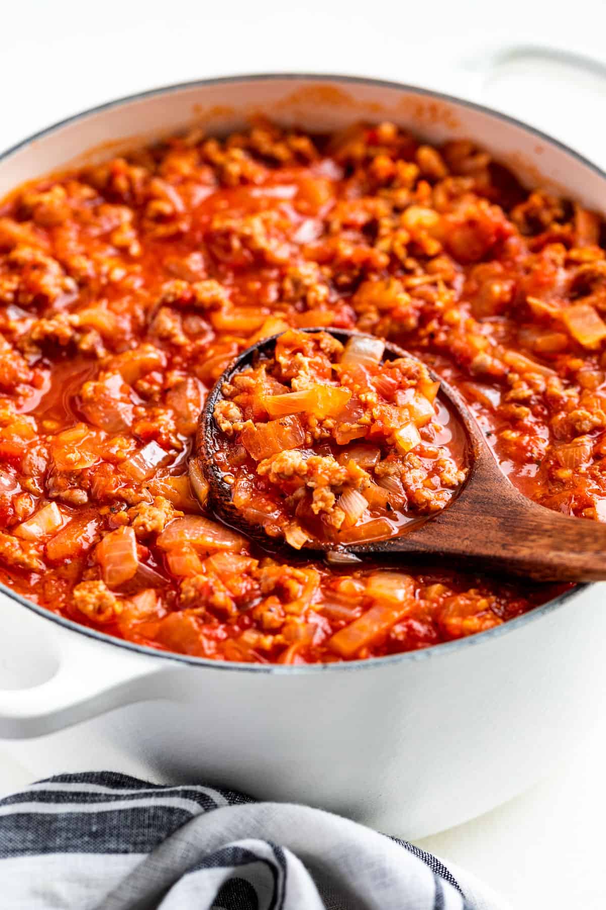 Side view of Homemade Spaghetti Sauce in a white pot with a wood ladle scooping some out.