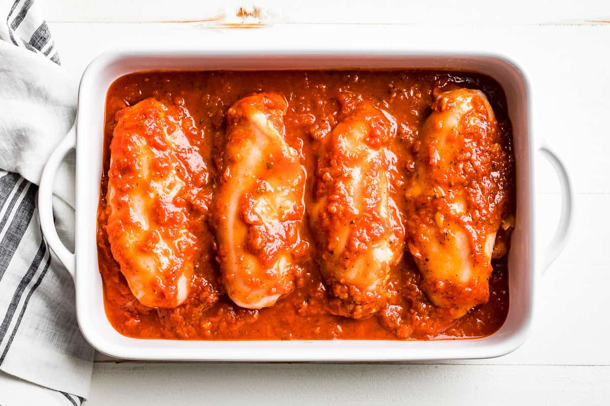 4 stuffed Pizza Chicken breasts in a white baking dish topped with tomato sauce.