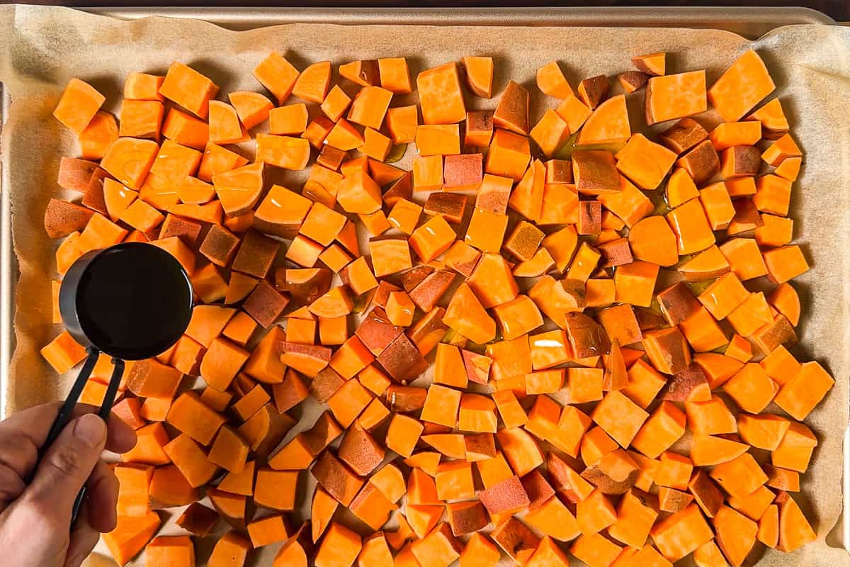 Pouring olive oil over cubed sweet potatoes on a parchment lined baking sheet.