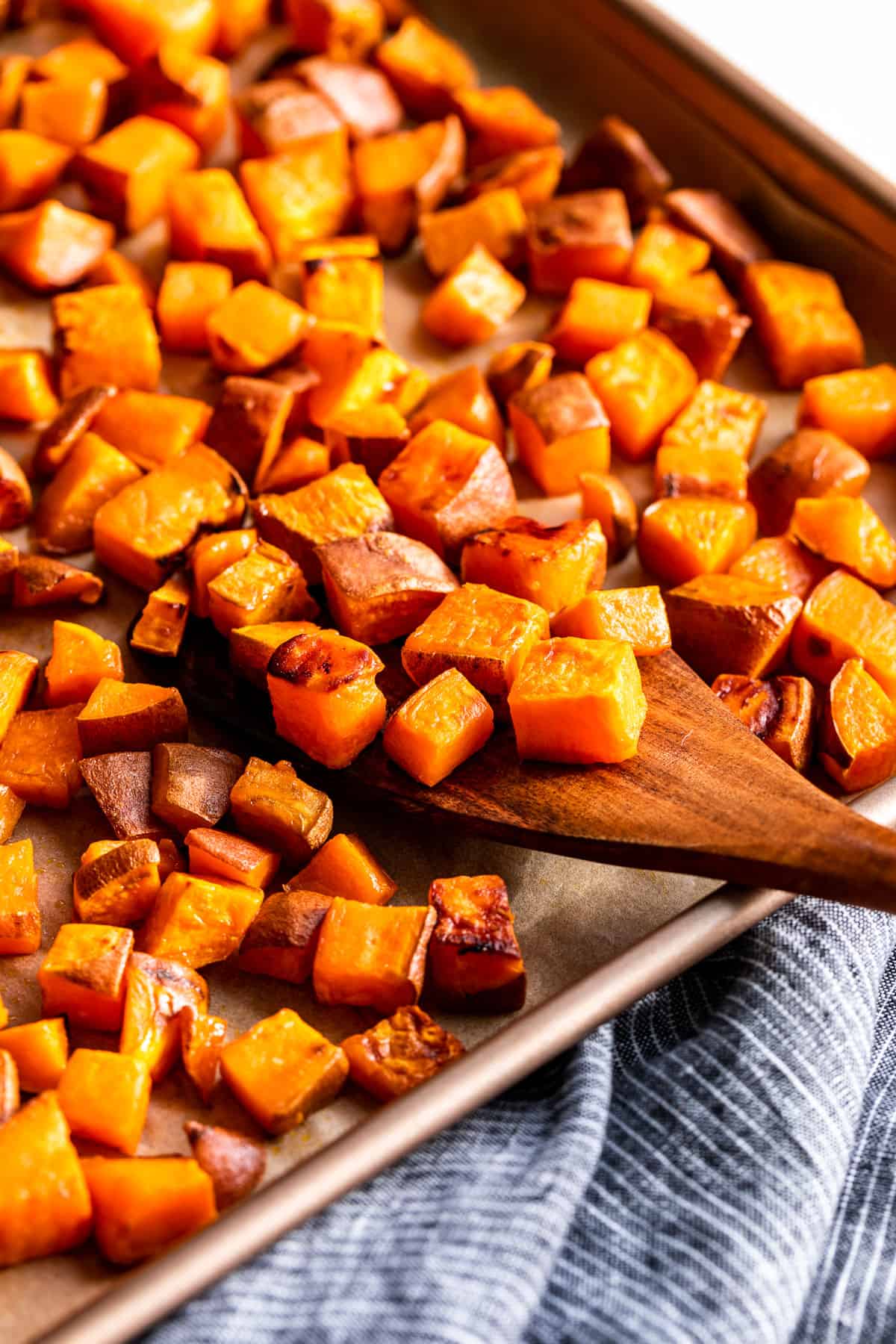 Side view of cubed Roasted Sweet Potatoes on a baking sheet with a wood spatula scooping some off.