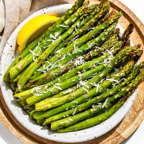 Finished air fryer asparagus on a pottery plate with a lemon wedge on the side sitting on a round wood plate.