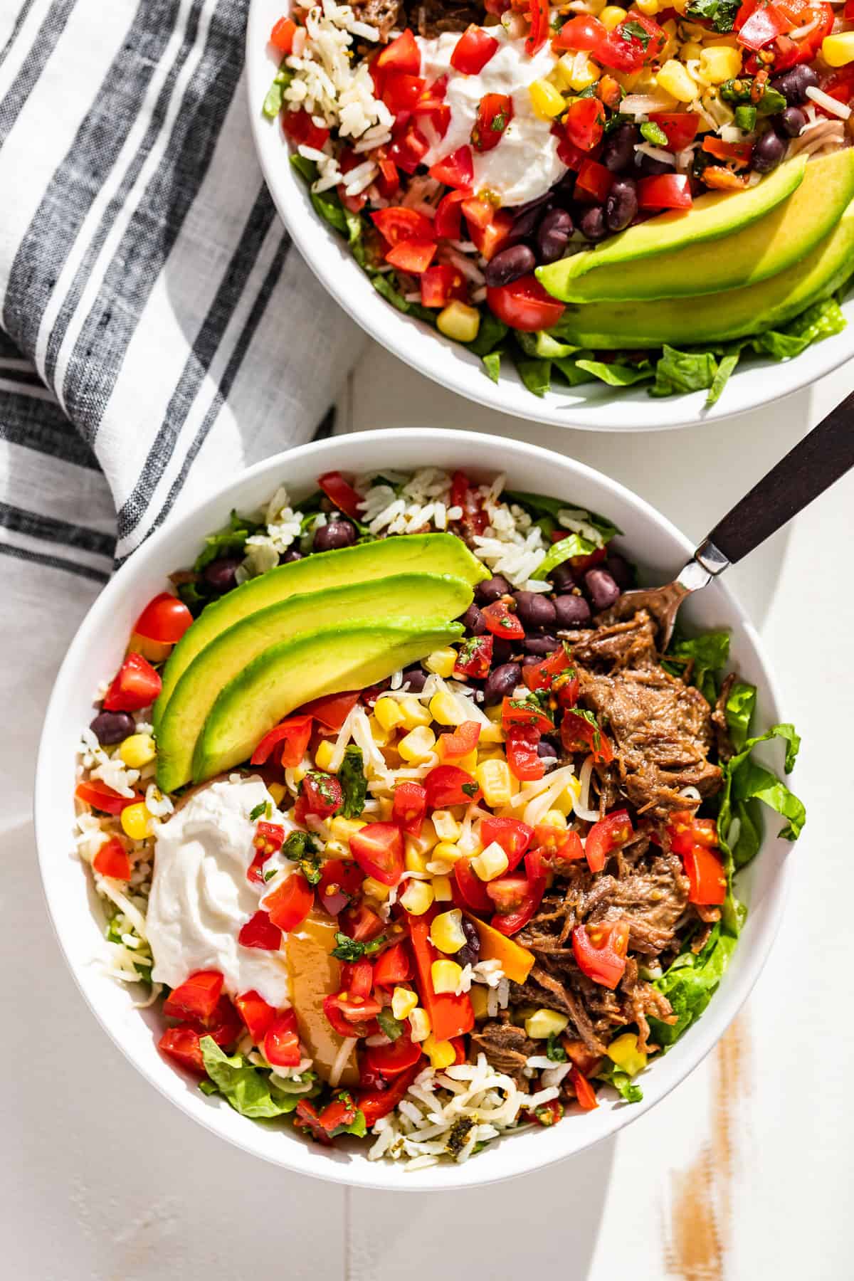 Straight down view of two Barbacoa Burrito Bowls topped with sliced avocado on a white background with a blue and white striped linen on the side.