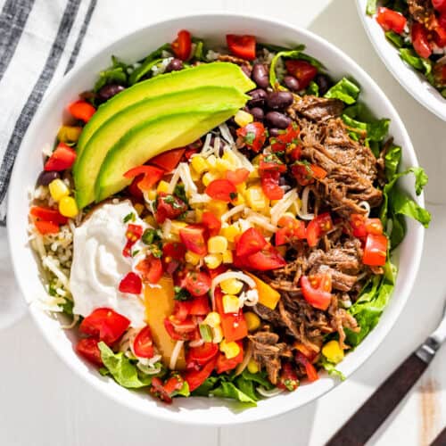 Finished Barbacao Burrito Bowls topped with sliced avocado on a white background with two forks on the side.