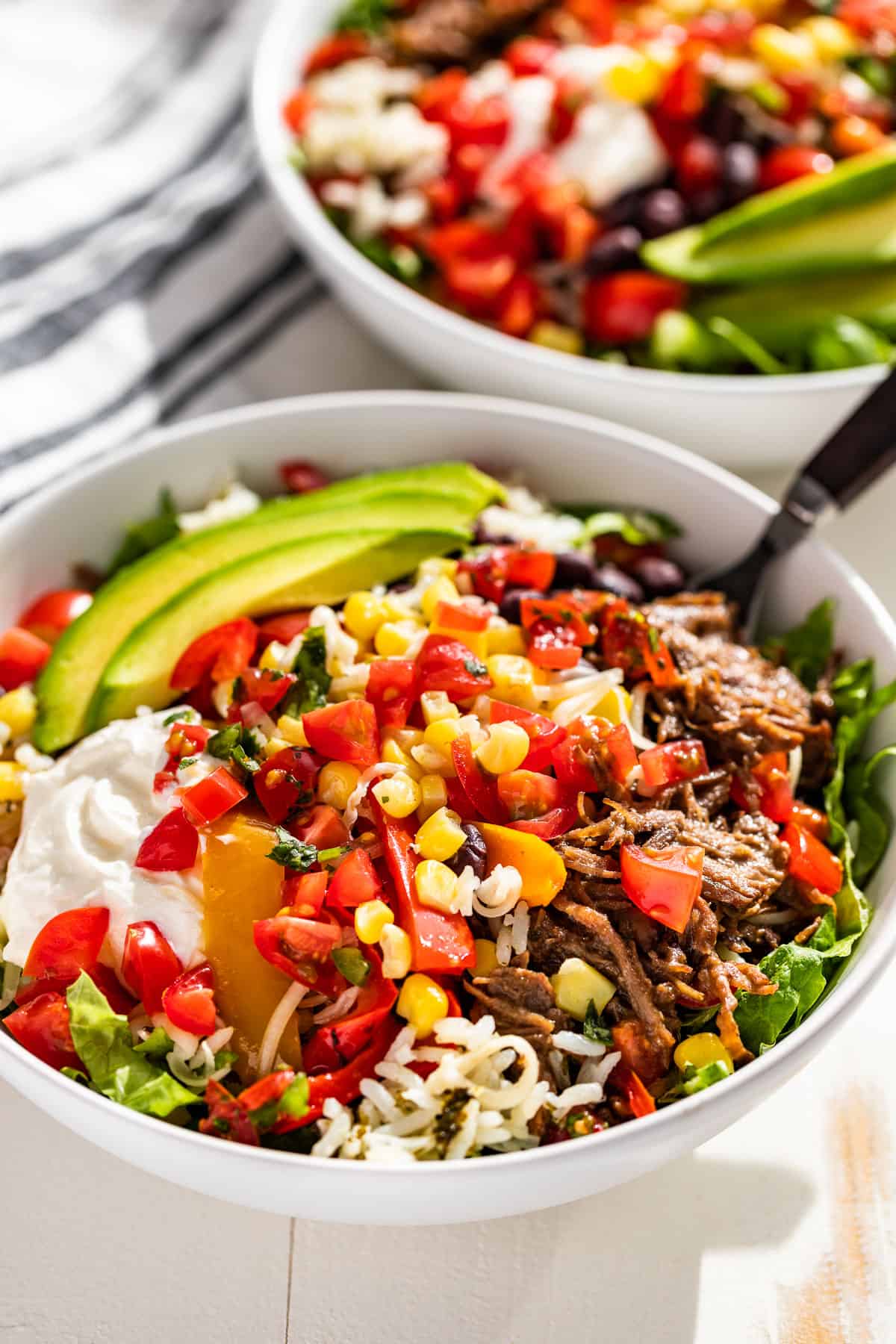 Side view of two Barbacoa Burrito Bowls topped with Pico de Gallo, sour cream and sliced avocado.