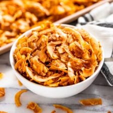 Coconut Chips in a white bowl with a cookie sheet of coconut chips in the background.