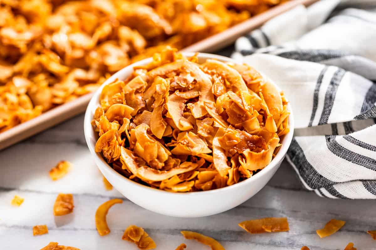 Coconut Chips in a white bowl with a cookie sheet of coconut chips in the background.