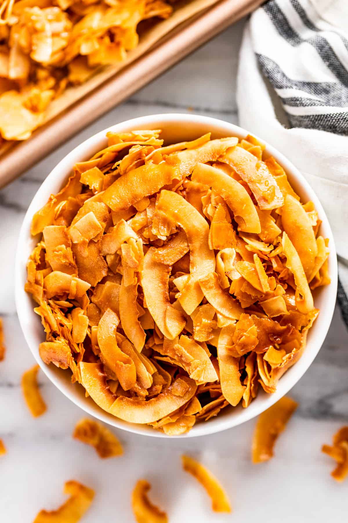 Straight down view of Coconut Chips in a white bowl with the cookie sheet of coconut chips on the side.