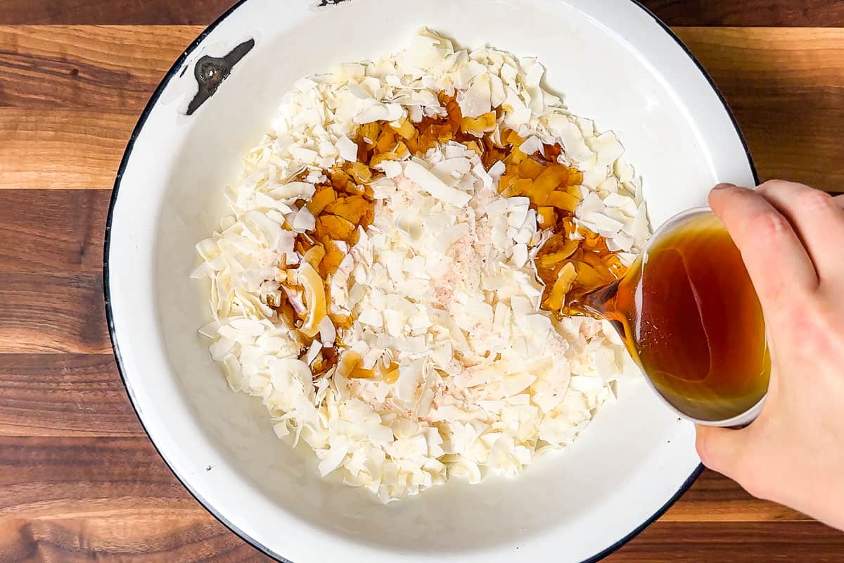 Adding maple syrup to coconut flake in a large white bowl.