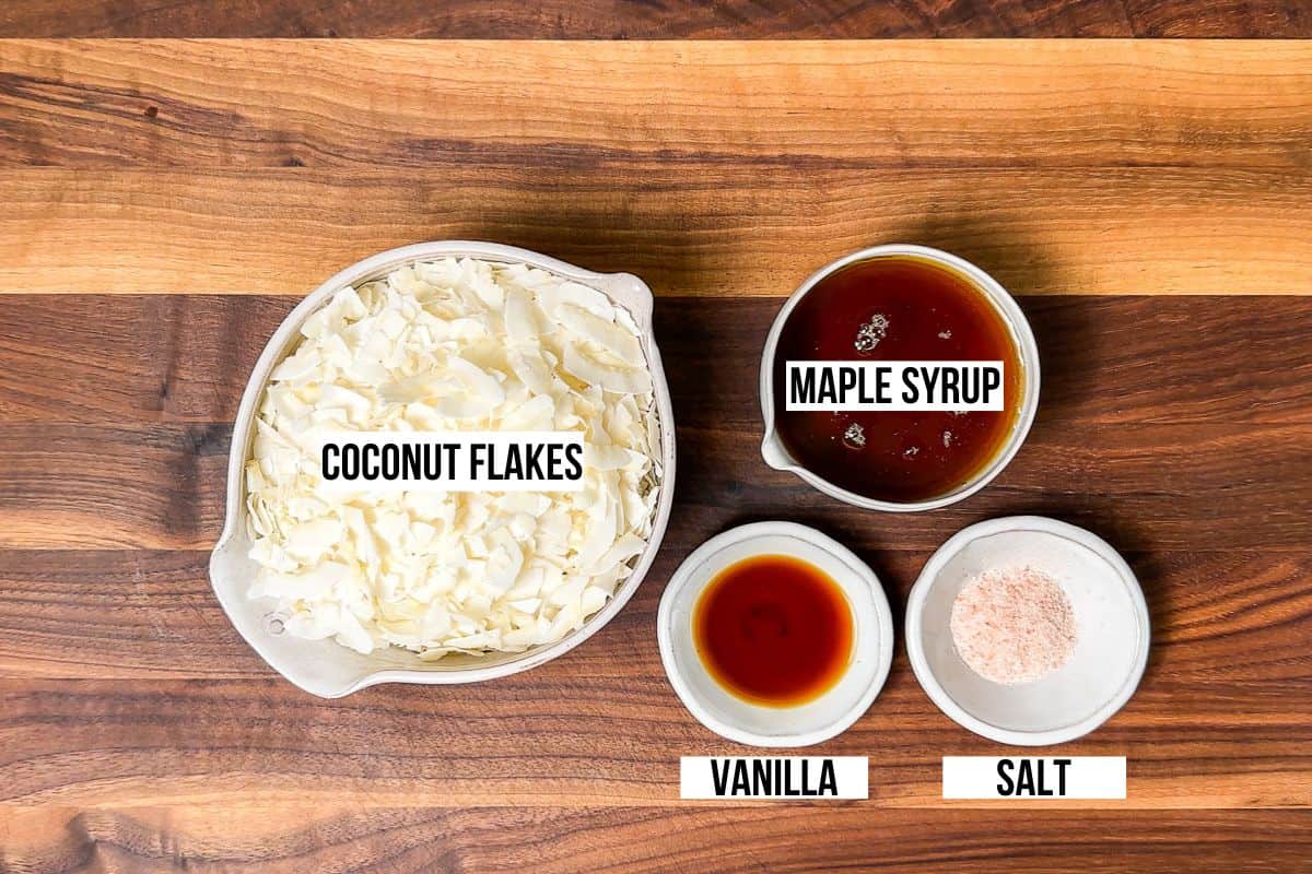 Coconut flakes, maple syrup, vanilla extract, and sea salt in bowls on a wood cutting board.