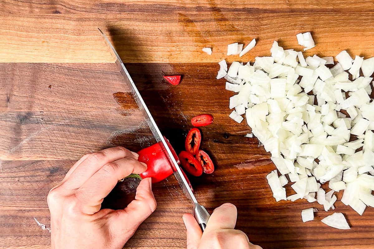 Diced onion and slicing a red chili on a wood cutting board with a chefs knife.