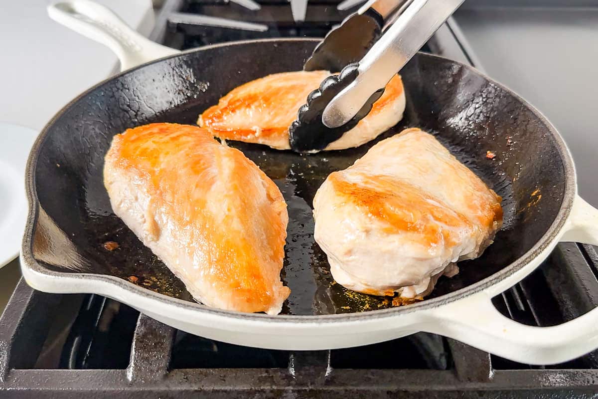 Browning the chicken breasts in a white enameled cast iron skillet and turn them with metal tongs.
