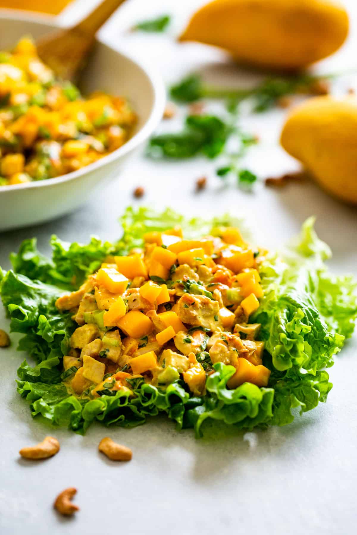 Curried chicken salad piled onto a large leaf of lettuce topped with diced mango and a bowl of chicken salad in the background.