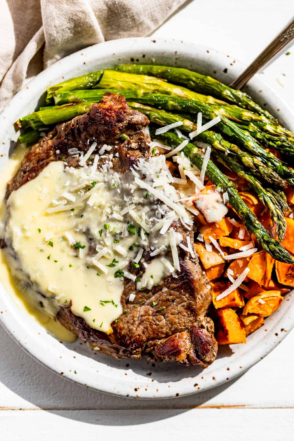 Garlic Parmesan Sauce drizzled over a ribeye steak with roasted sweet potatoes and asparagus on the side sprinkled with parmesan.